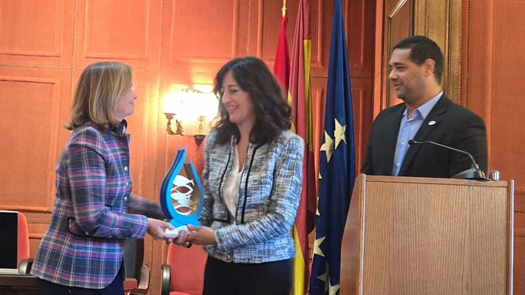 En la imagen se observan tres personas, dos mujeres y un hombre. Una de las mujeres, rubia y vestida de rojo entrega el premio Jacumar a otra mujer de mediana edad, morena y vestida de azul claro. El hombre permanece en segundo plano observando la escena.